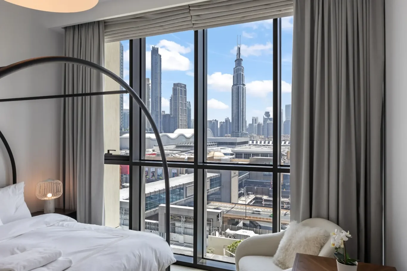 Hotel bedroom with white bedding and a curved floor lamp, large windows framing a city skyline outside.