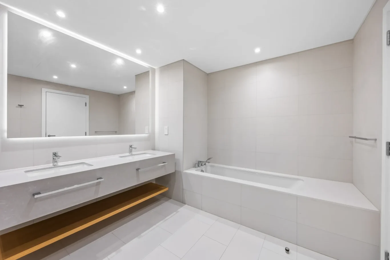 Modern white bathroom with a double sink vanity, large backlit mirror, and a built-in wooden shelf beneath the sink.