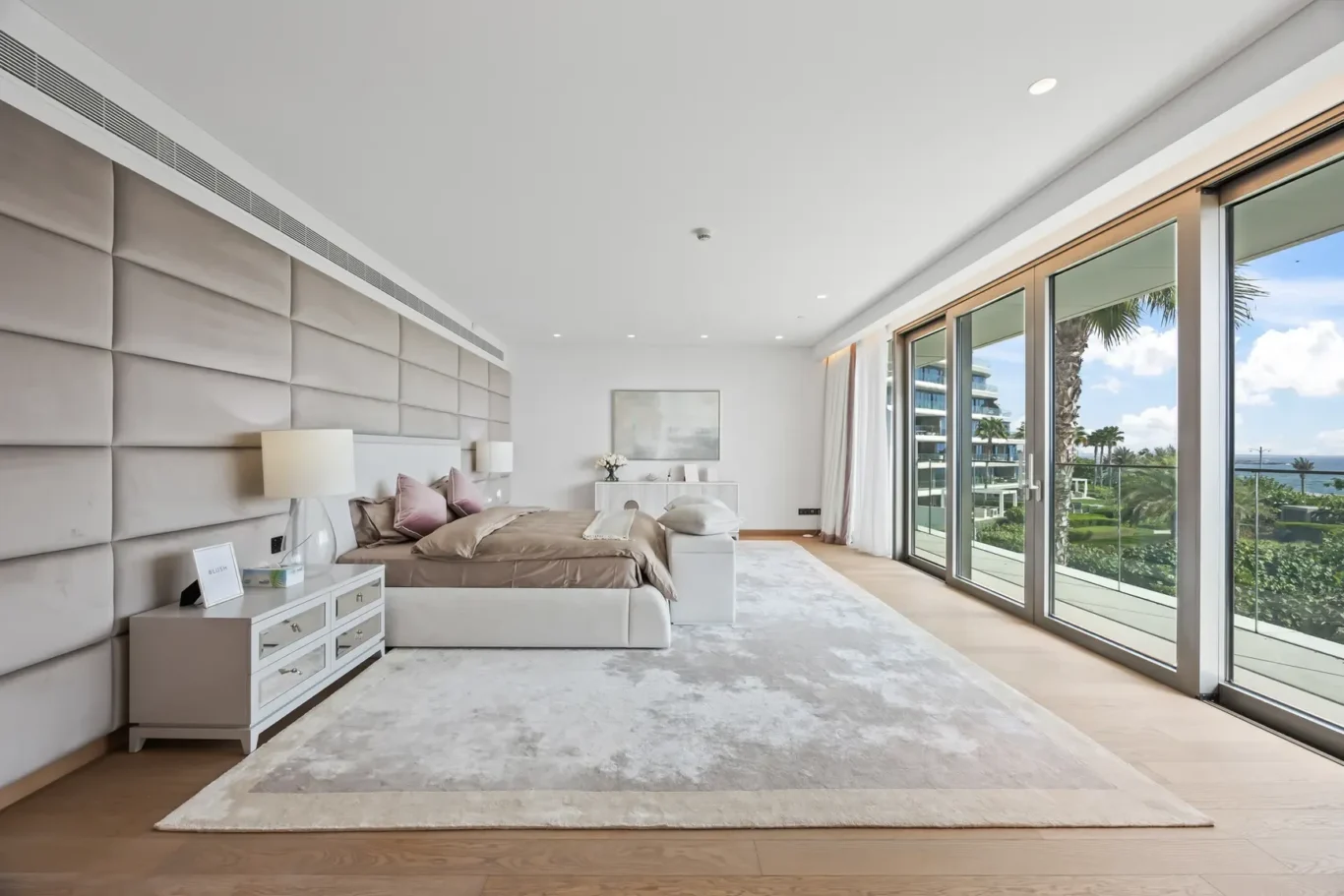 Luxury hotel bedroom with a tall beige tufted headboard, white nightstands, and neutral bedding, plus a view of a balcony and ocean beyond the glass walls.