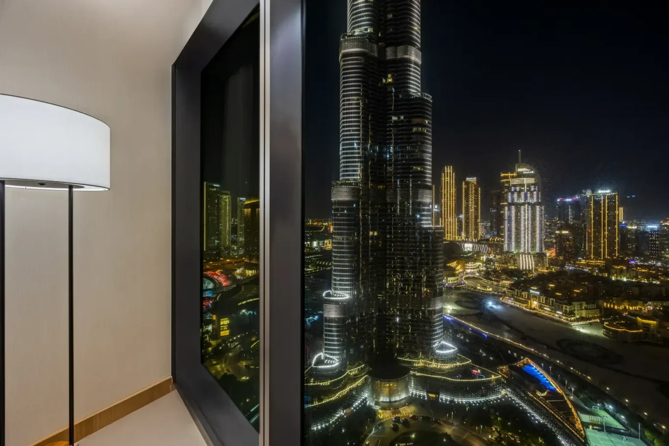 Nighttime city skyline viewed from a high-rise hotel room window, with a tall illuminated skyscraper dominating the scene in the city lights.