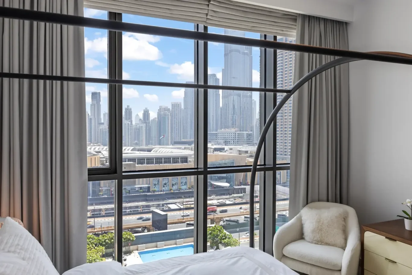 Hotel room with a large window and a city skyline view; bed in the foreground and a white armchair by a wooden desk on the side.