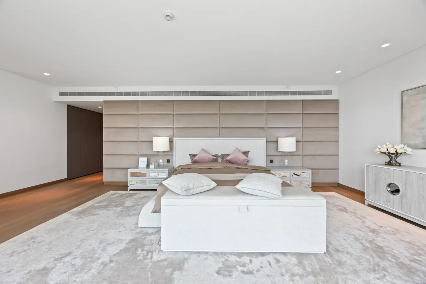 Luxurious bedroom with a tall padded beige headboard, king bed, matching nightstands and lamps, and a light gray rug.