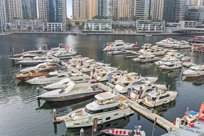 Busy marina with numerous white yachts docked along floating piers and a city skyline in the background.