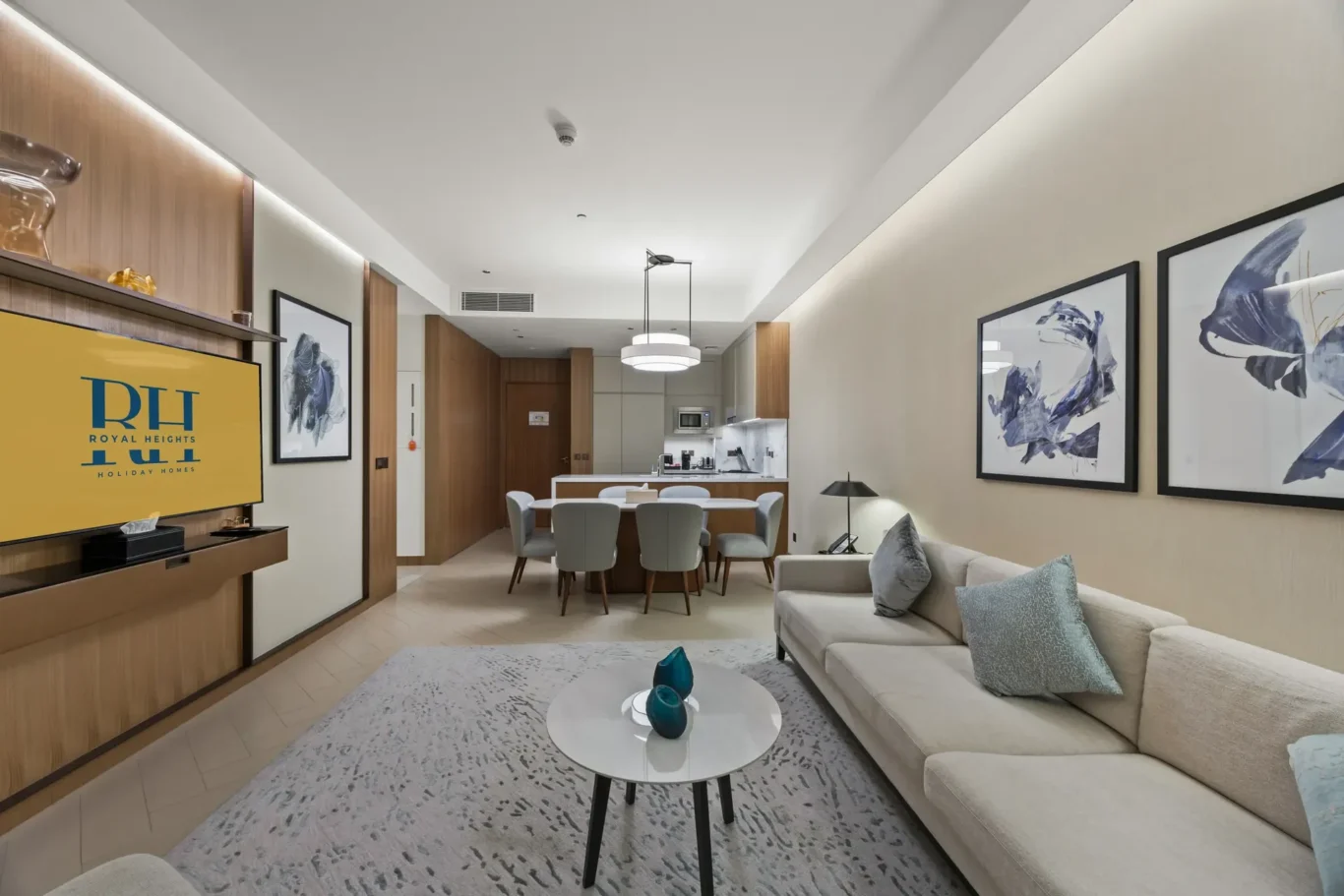 Open-plan hotel living area with a beige sectional sofa, round coffee tables, gray rug, and a kitchen in the background.