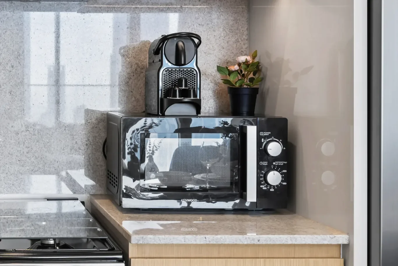 Kenwood microwave on a beige countertop with a compact coffee machine on top and a small potted plant beside it in a modern kitchen.