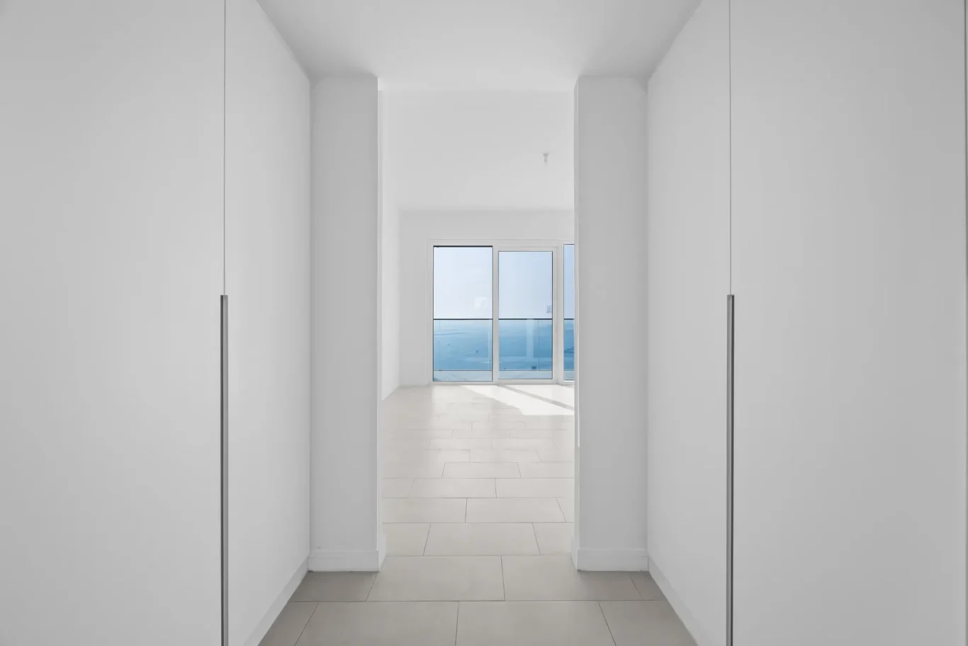 Bright white hallway with built-in wardrobes on both sides, opening to a glass door revealing a balcony and blue sea beyond.