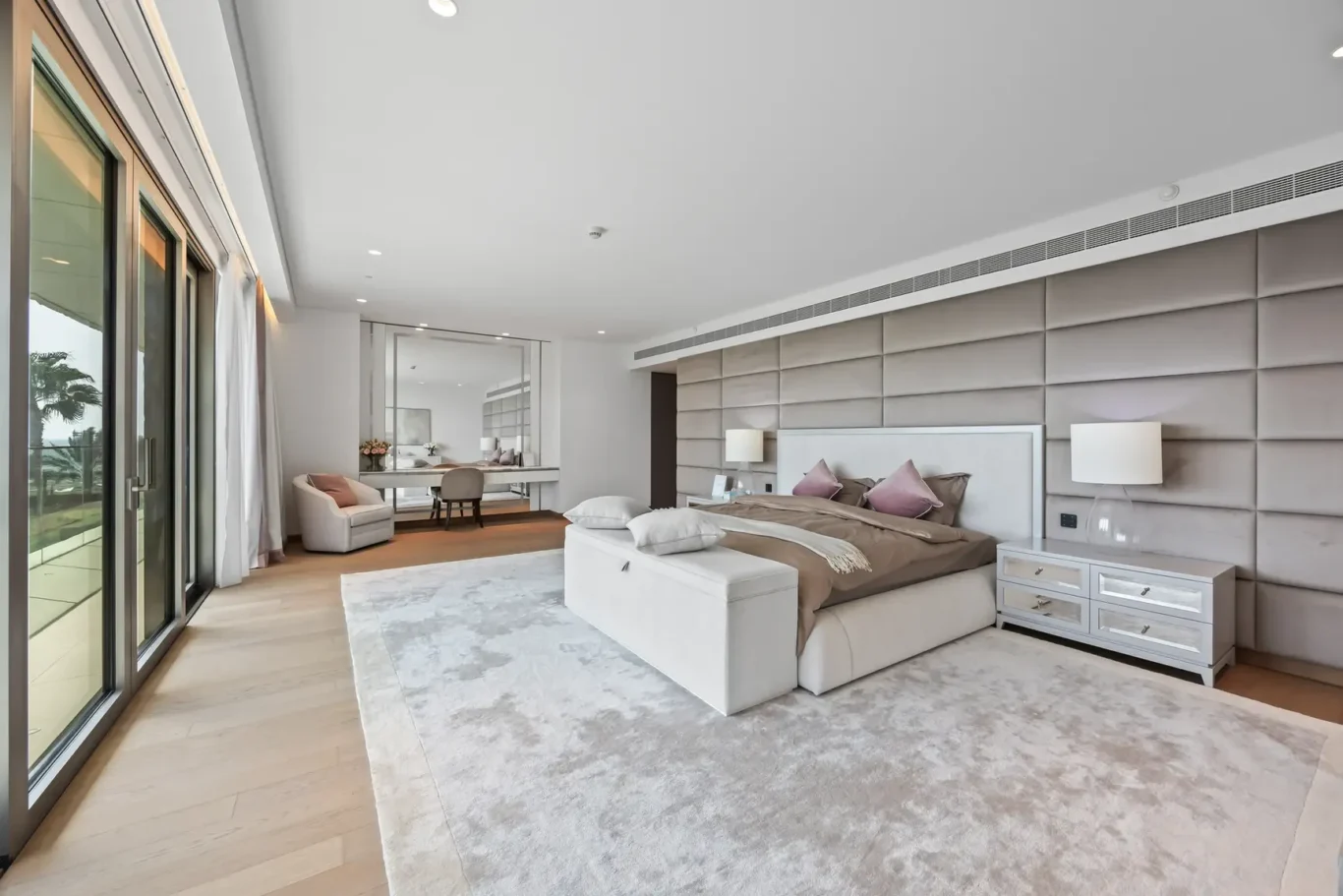 Bright modern bedroom with a large padded headboard wall, white bed, beige bedding, and two mirrored nightstands in a neutral palette.