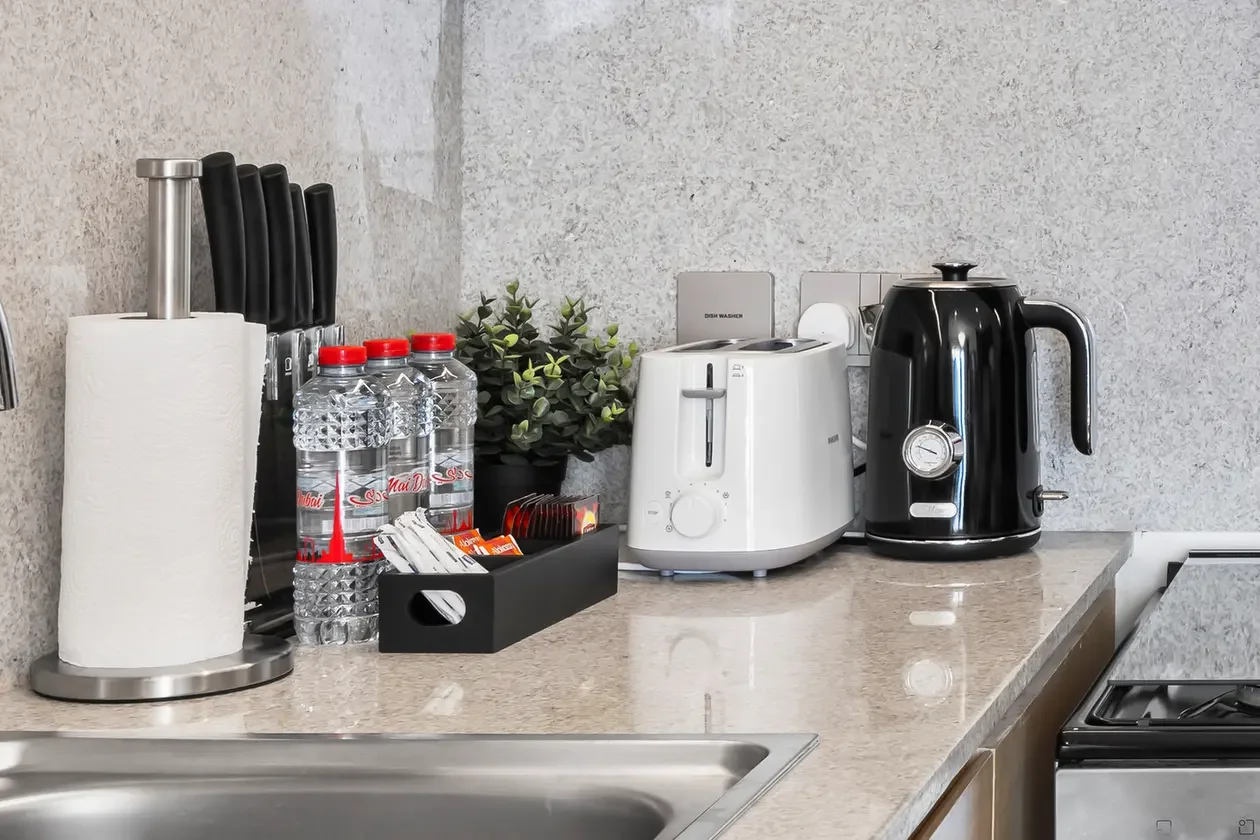 White toaster and black electric kettle sit on a marble kitchen counter with a paper towel roll, bottled water, knife block, and a small plant nearby.