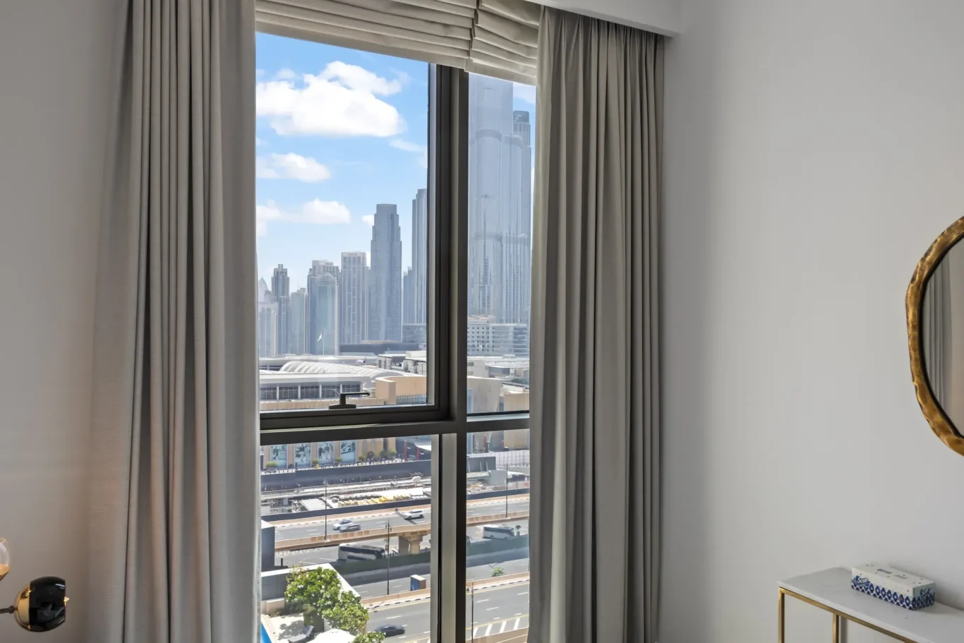 Hotel room window with beige curtains, overlooking a city with tall skyscrapers and clear blue sky.