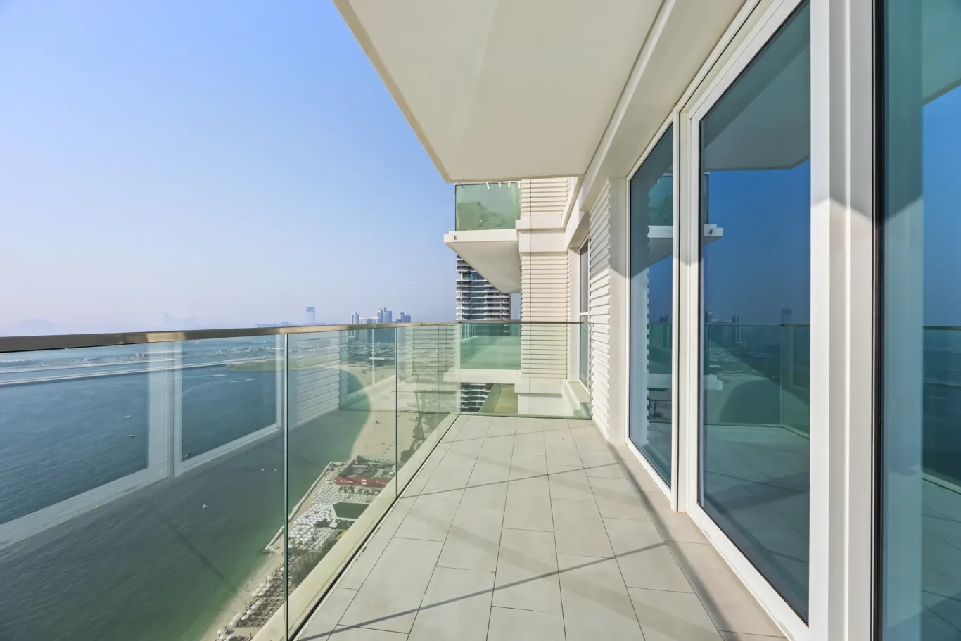 Modern balcony with glass railing overlooking the ocean and distant city skyline, adjacent to a white building with large glass doors.