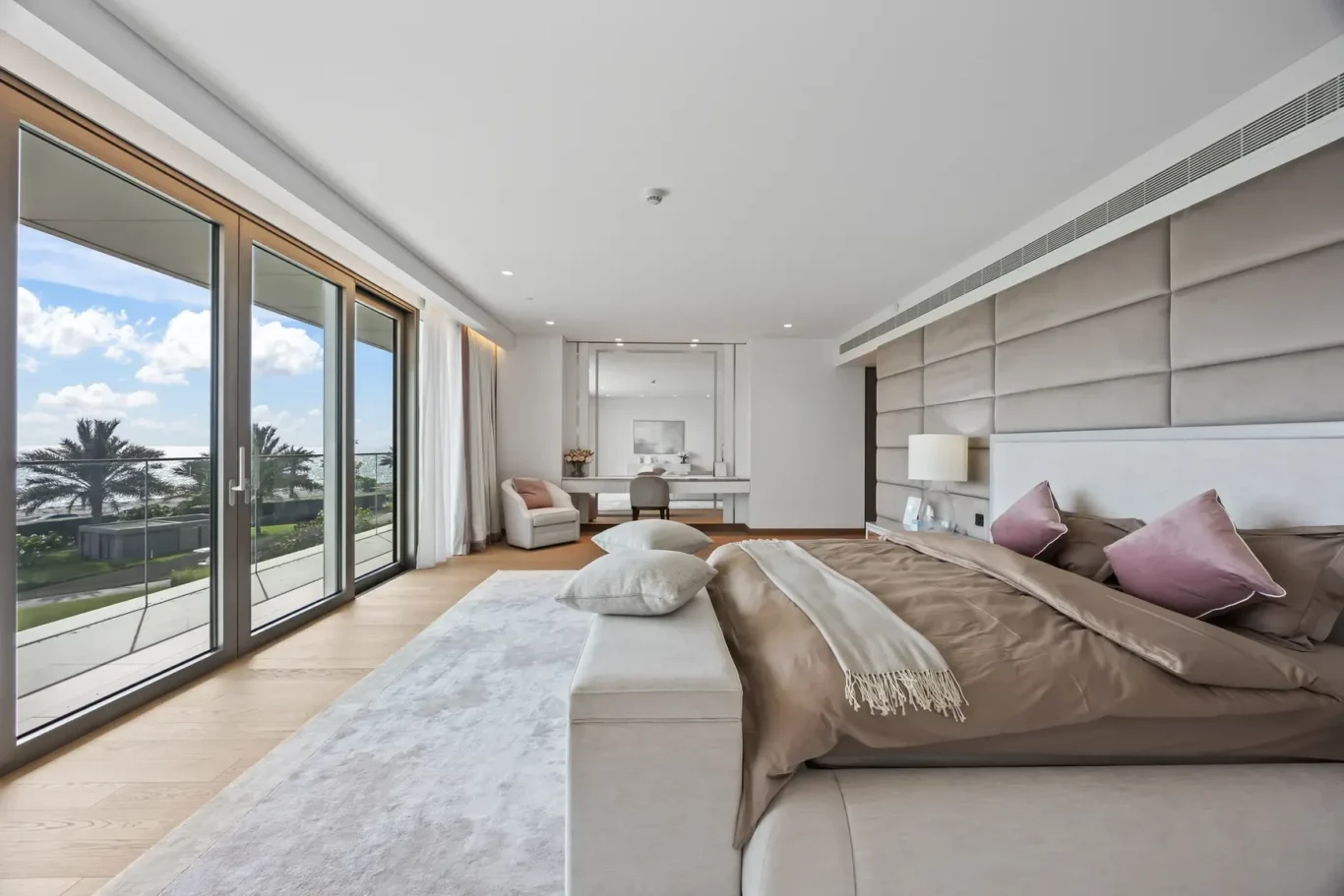 Luxurious hotel bedroom with a king bed, soft beige bedding and a light gray headboard, plus a balcony view of palm trees and the ocean outside.