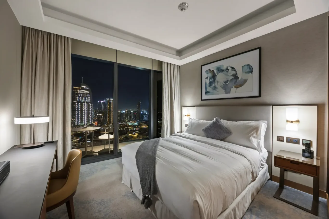 Hotel bedroom at night with a large bed, side tables, abstract artwork, and a city skyline visible through floor-to-ceiling windows.
