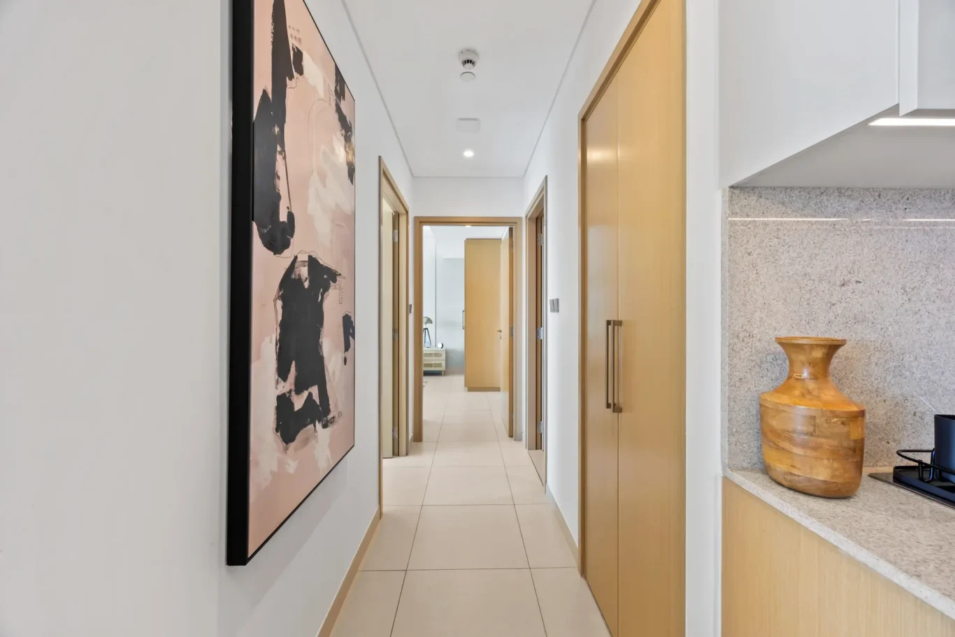 Bright modern hallway with white walls, beige wooden doors, and a large abstract painting on the left; granite counter and a wooden vase on the right.