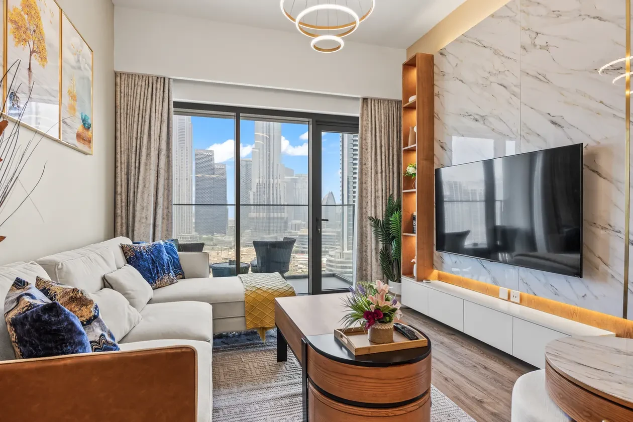 Modern living room with a white sectional sofa, colorful pillows, and a wooden coffee table, overlooking a city skyline through glass doors.