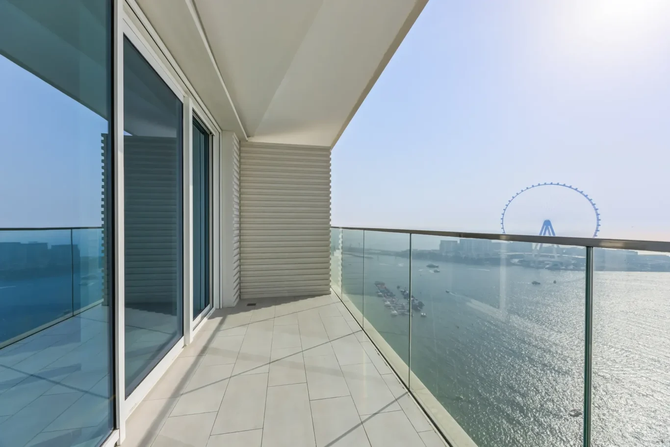 Balcony with glass railing and sliding glass doors, overlooking a harbor and a distant Ferris wheel.