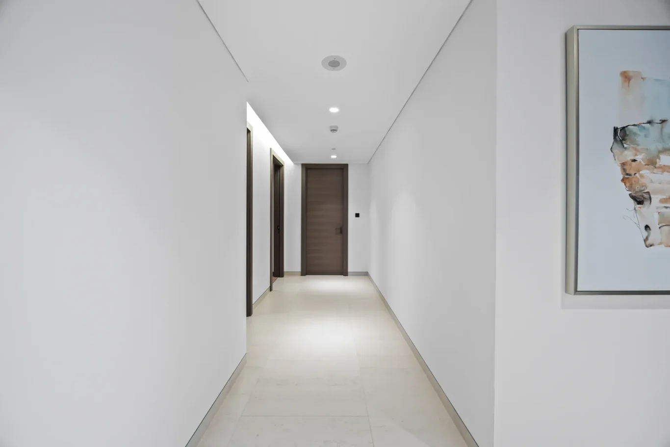 Long, bright hallway with wooden doors on the left and abstract artwork on the right; a closed wooden door at the end of the corridor.
