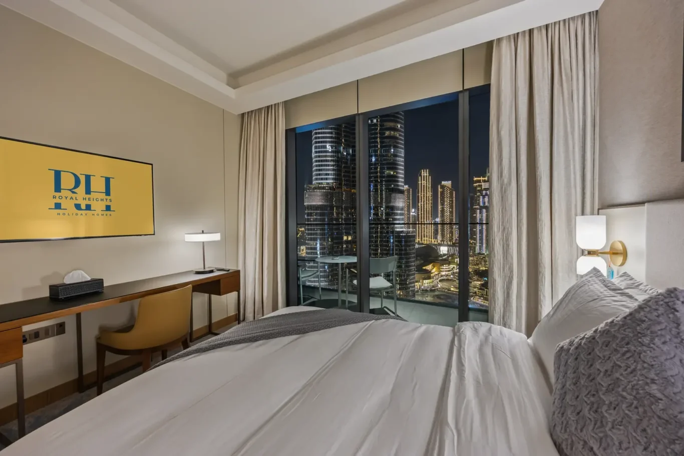 Hotel bedroom at night with city skyline visible through large windows; bed in foreground, desk and chair on the left, beige curtains.