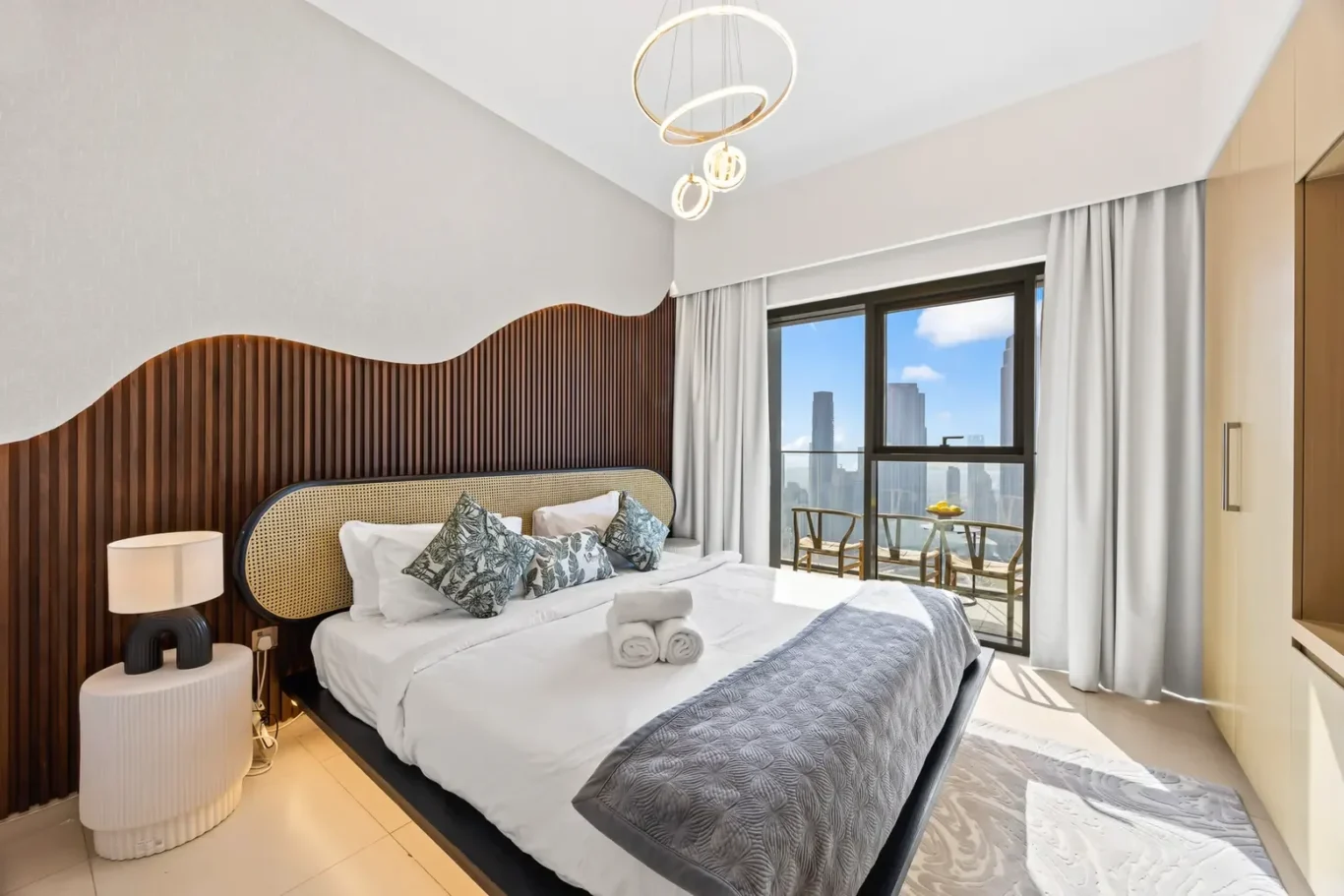 Hotel bedroom with a curved wooden slat headboard, white bedding, decorative pillows, and folded towels on the bed; large window opens to a balcony with a city skyline view.