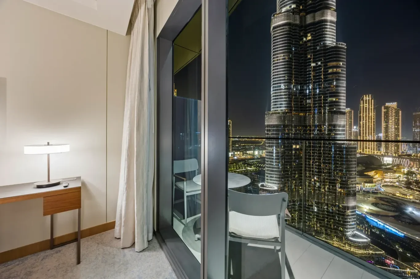 Hotel room interior at night with a desk and lamp, and a glass balcony door opening to a illuminated city skyline outside.