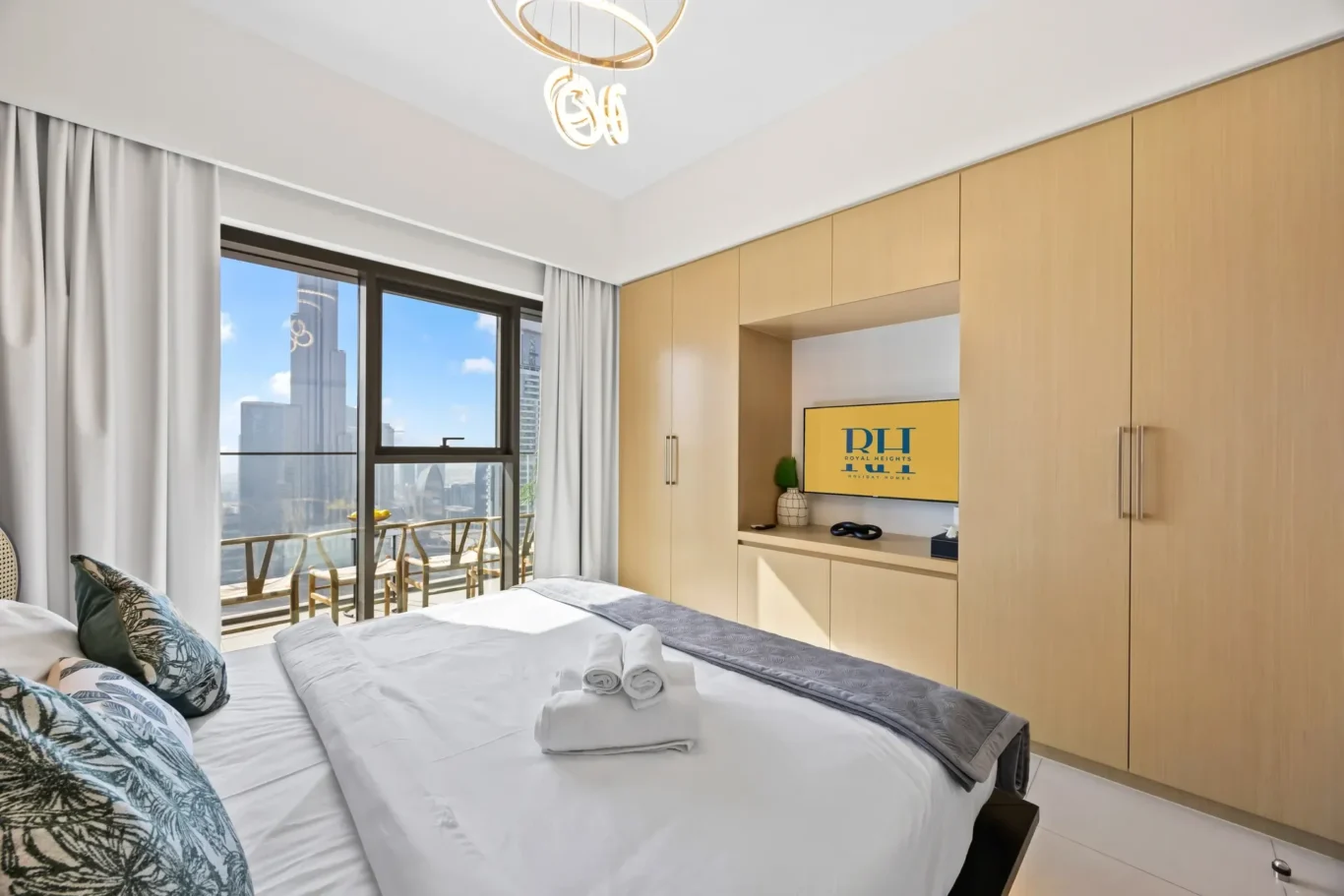 Contemporary hotel bedroom with a king bed, white linens and decorative pillows, and built-in light-wood wardrobes surrounding a TV nook. A balcony and city skyline are visible through the large window.