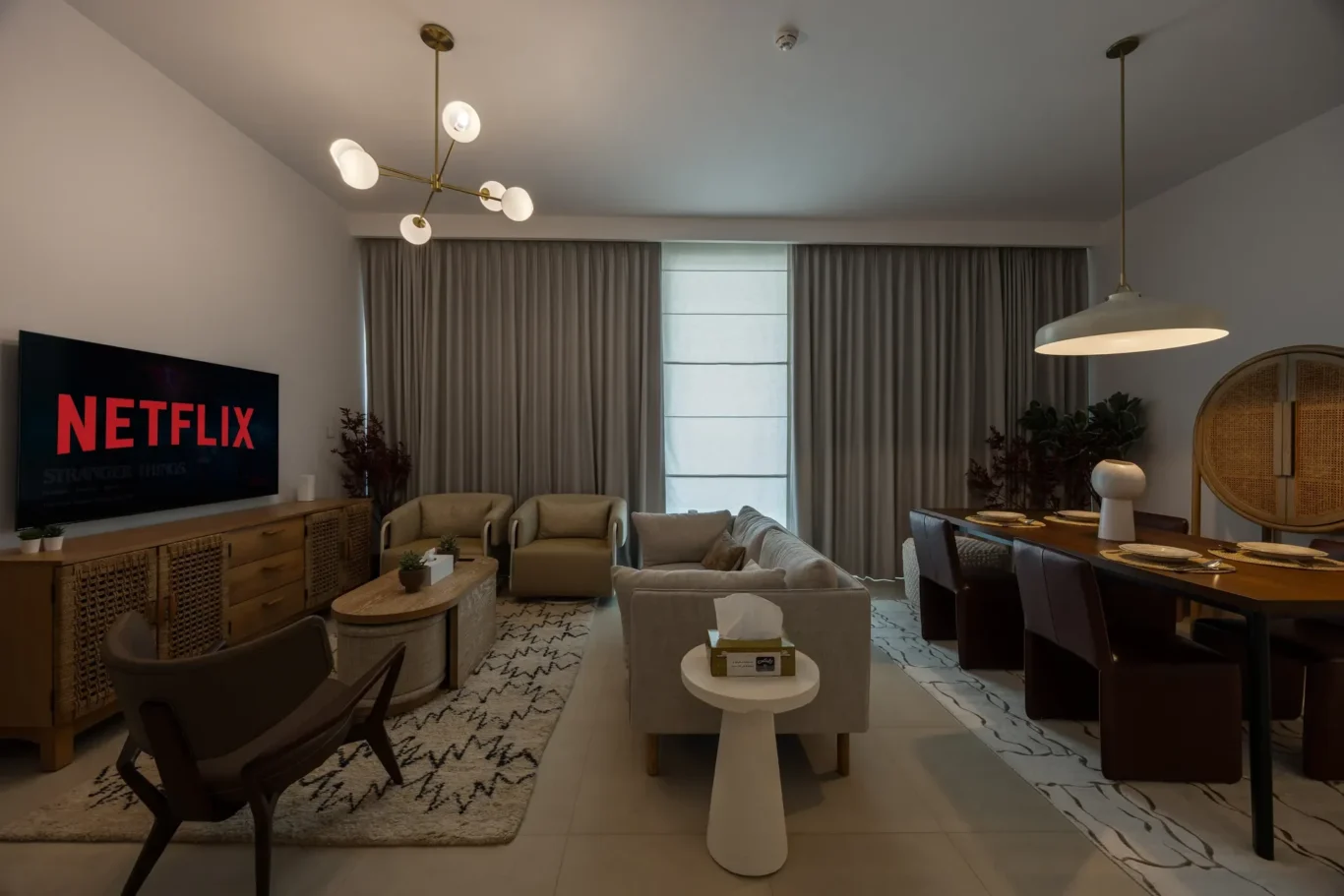 Living room with a wall-mounted TV displaying Netflix, beige sofas, and a wooden credenza under a modern chandelier.