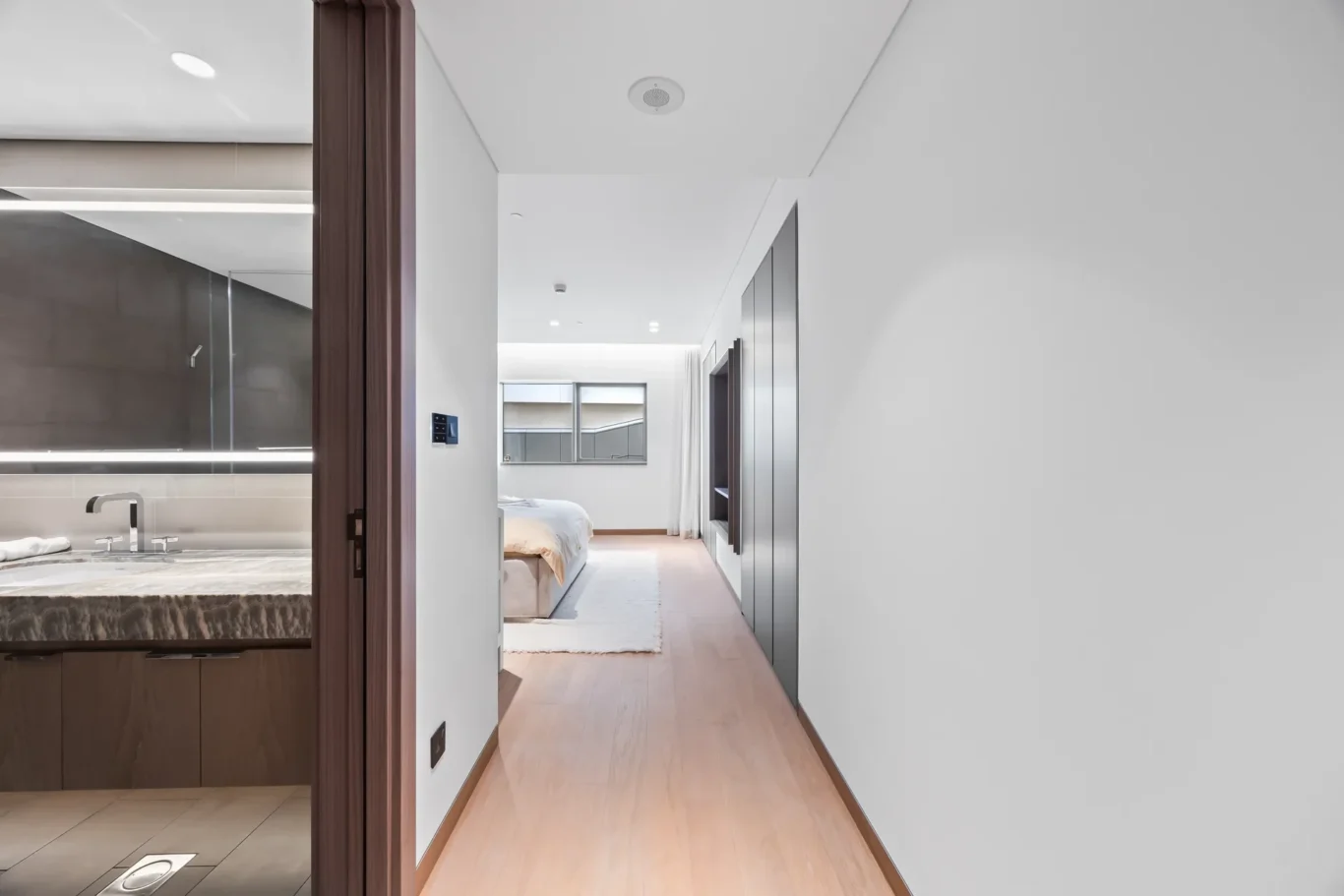 Bright modern hallway leading to a bedroom with a neatly made bed and full-height wardrobes on the right; bathroom area visible to the left.