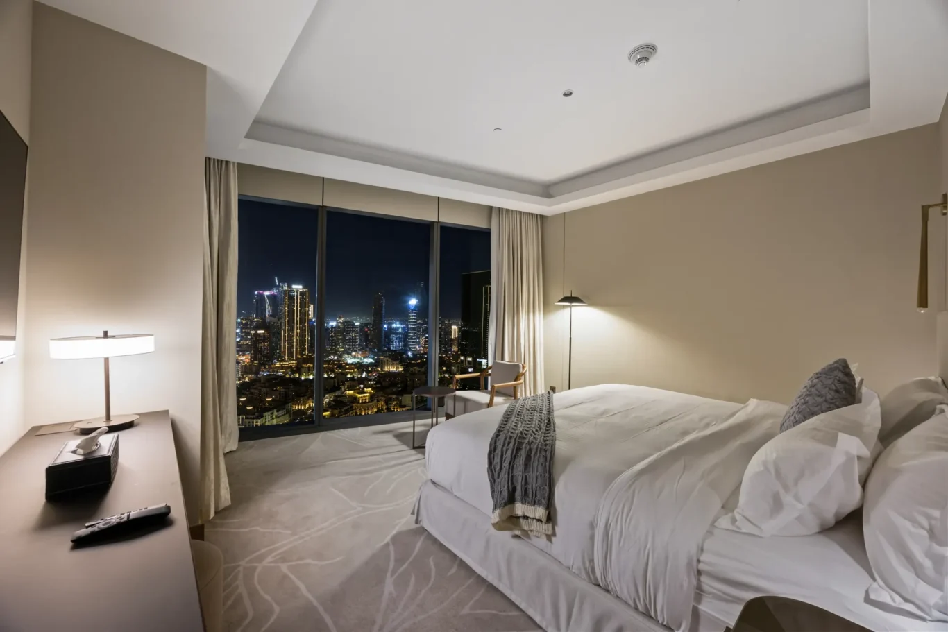 Hotel suite with two white beds facing a city skyline at night through large windows.