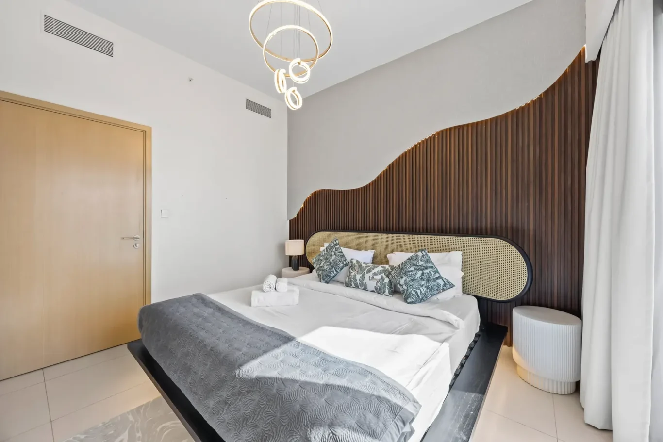 Modern hotel bedroom with a curved wooden slat headboard, circular pendant chandelier, and a king bed with botanical-pattern pillows.