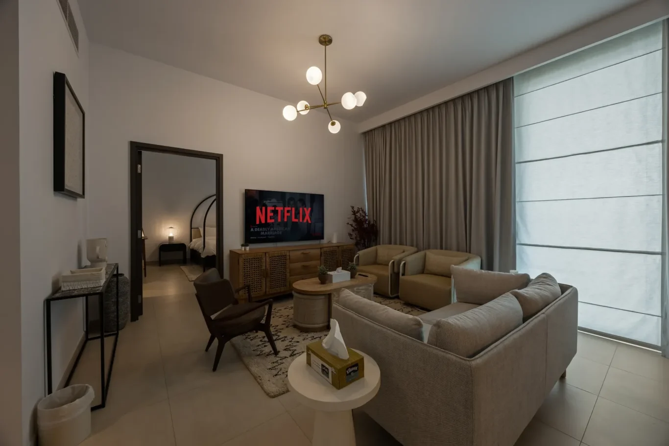 Living room with beige sofas, a round coffee table, and a TV showing Netflix; large window with light curtains on the right.