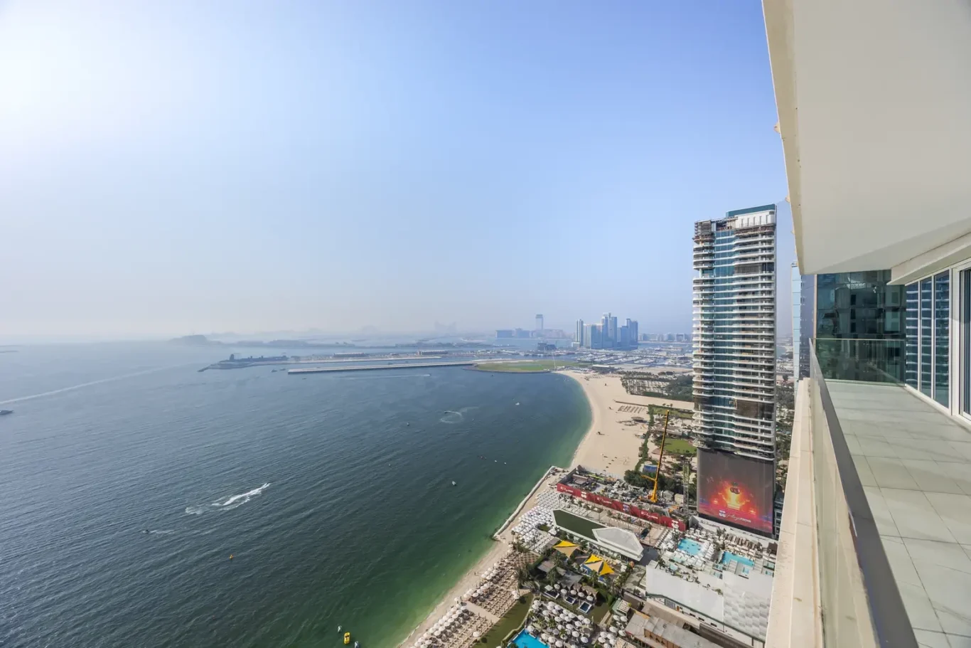 Aerial view of a sandy curved beach beside a calm blue bay, with modern high-rise buildings along the coast and a distant city skyline.