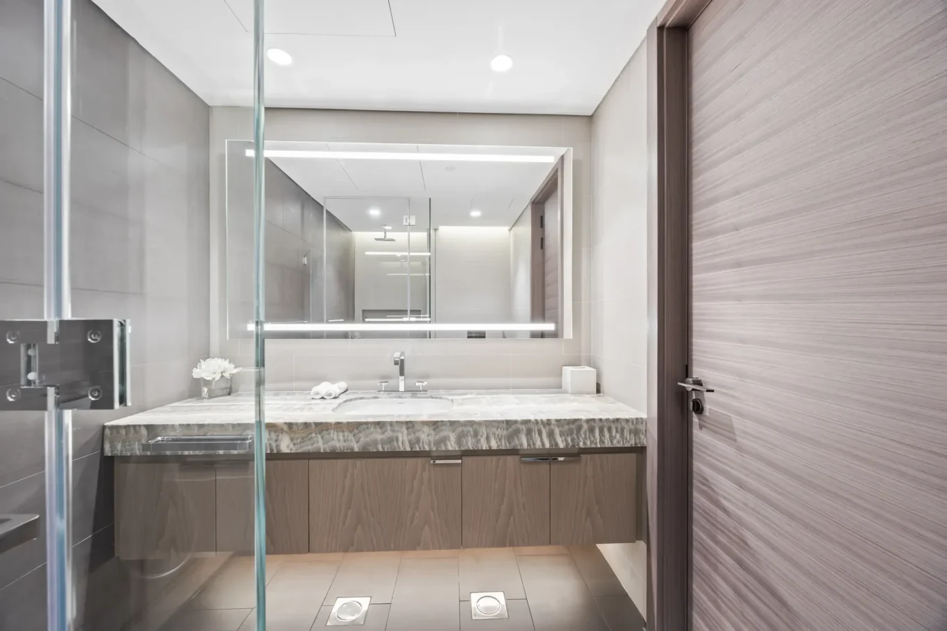 Modern bathroom featuring a marble countertop with an undermount sink, a large backlit mirror, and a glass shower enclosure; grey tiled walls and wood-tone cabinetry beneath the sink.