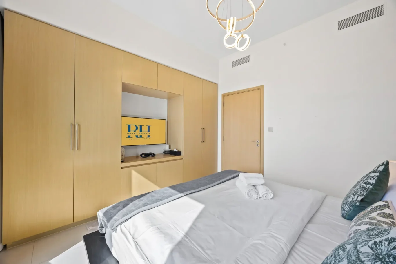 Bright hotel bedroom with a wooden wardrobe wall, recessed TV, and a neatly made bed with towels in the foreground.