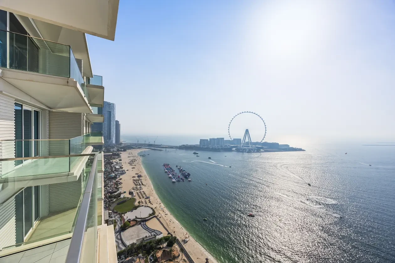 View from a high-rise balcony overlooking a sandy beach, calm blue water, and a distant Ferris wheel (Ain Dubai) by the coast on a sunny day.