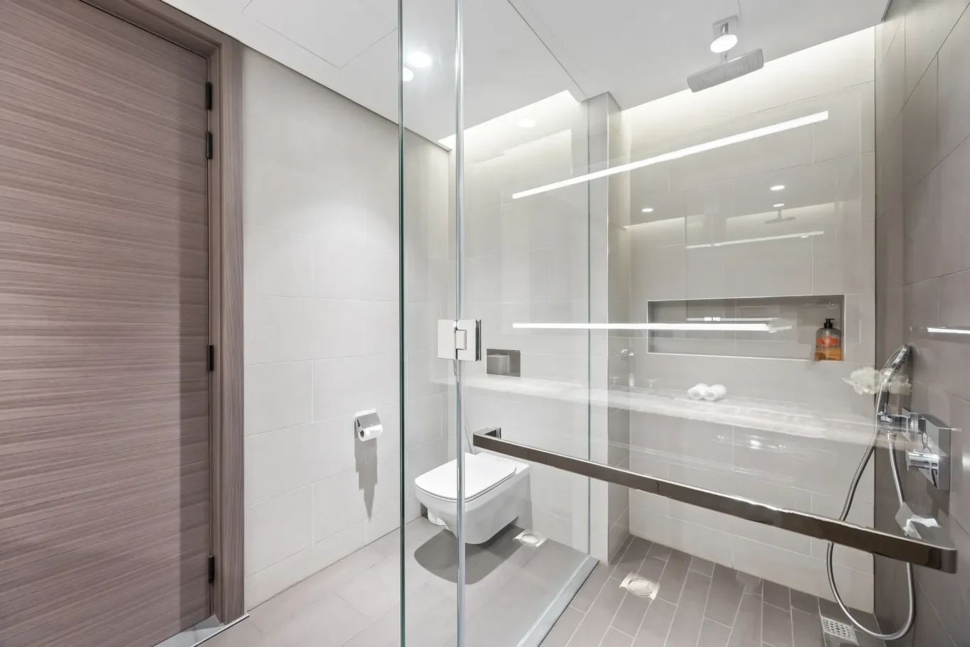 Modern bathroom with a glass shower enclosure, wall-hung toilet, and a wooden door on the left.