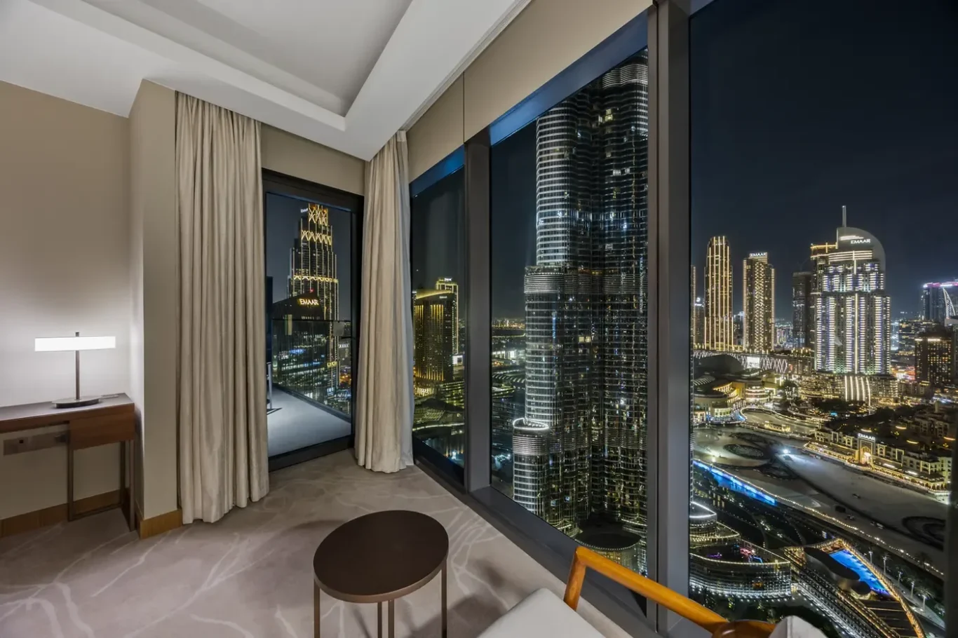 Hotel room at night with floor-to-ceiling windows showing a brightly lit city skyline of skyscrapers and streets below far outside the glass doors.