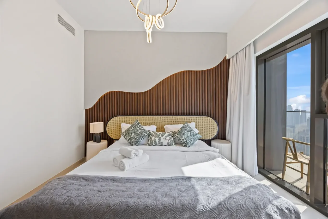 Modern hotel bedroom with a wave-shaped wooden headboard and rattan panel behind the bed, white bedding and patterned throw pillows.