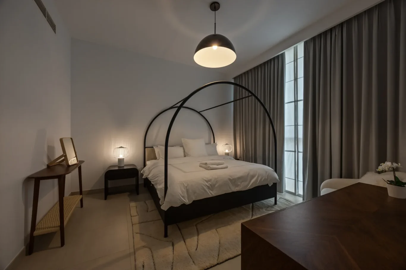 Modern hotel bedroom with a white bed, dark circular-arch metal frame, two bedside lamps, and gray curtains