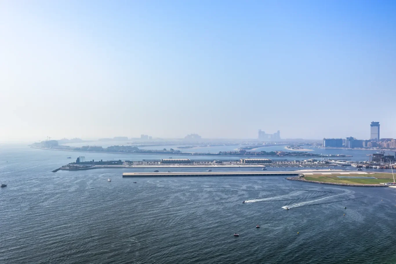 Coastal harbor with breakwaters and calm blue water under a clear sky, distant city skyline on the horizon