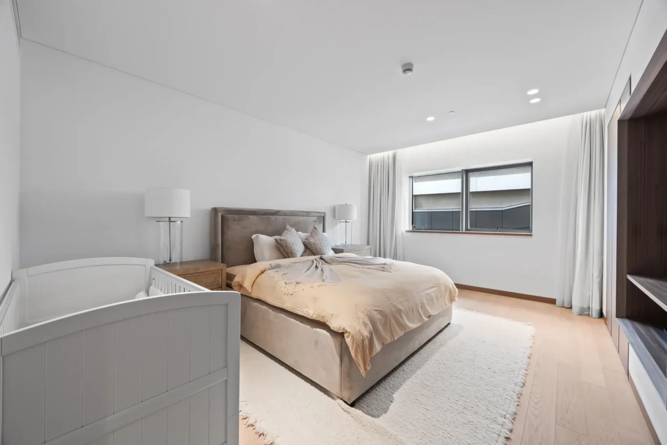 Modern bedroom with a large bed, tan upholstery, nightstands, and a white crib in the foreground; neutral decor and soft lighting.