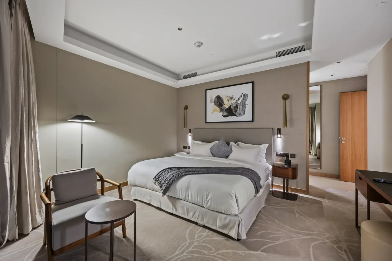 Hotel bedroom with king bed, gray headboard, two nightstands, framed artwork, and a seating area by the window.