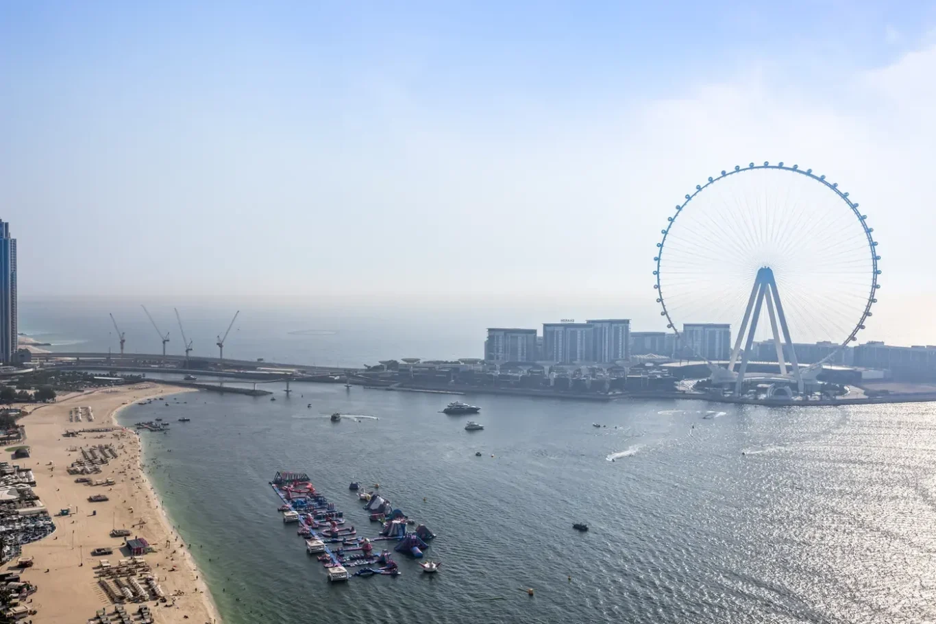 Aerial view of Dubai Marina with the Ain Dubai Ferris wheel, beach, and boats on calm water on a clear day