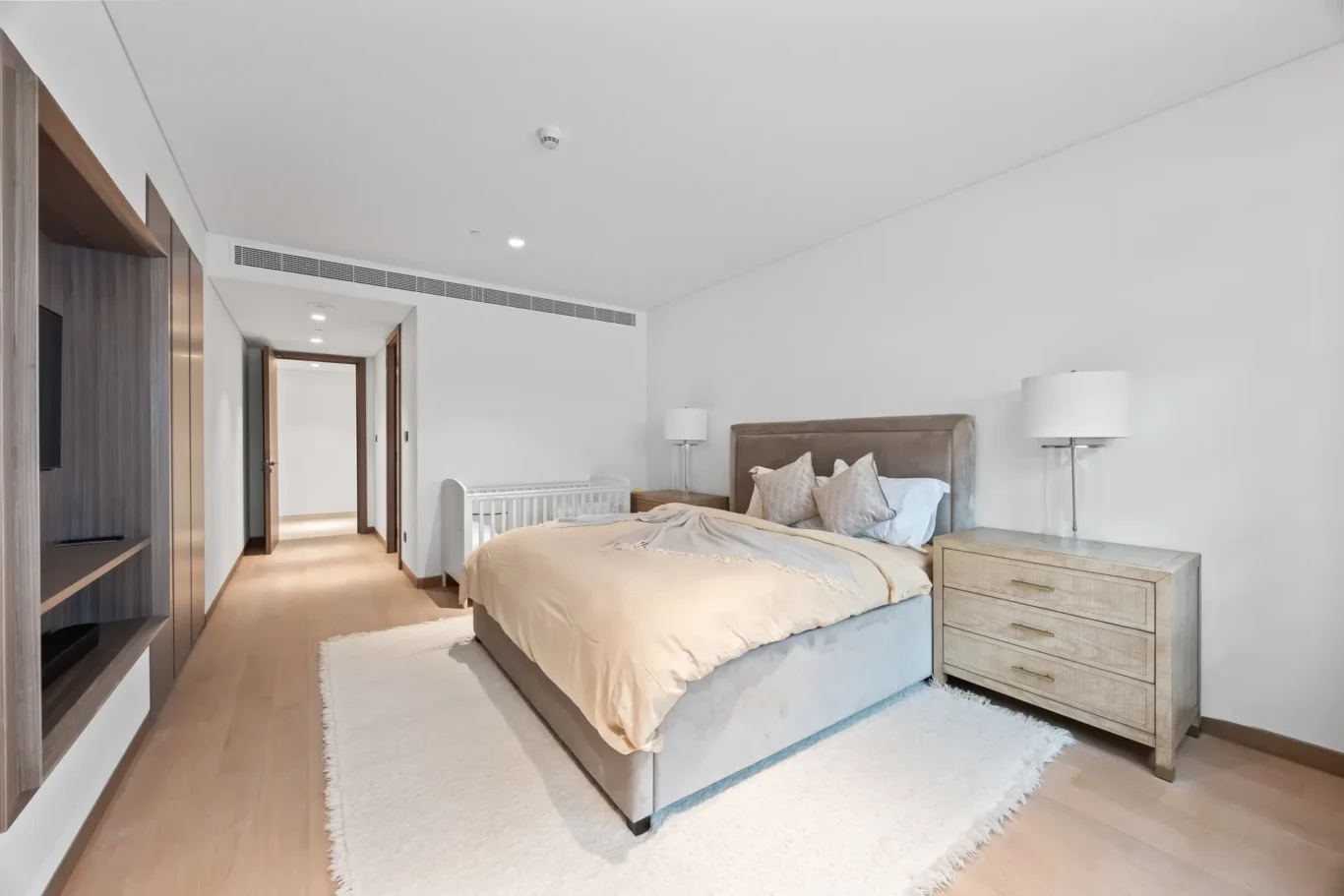 Bright bedroom with a beige upholstered bed, two nightstands with lamps, a white shag rug, and a crib along the wall to the left.