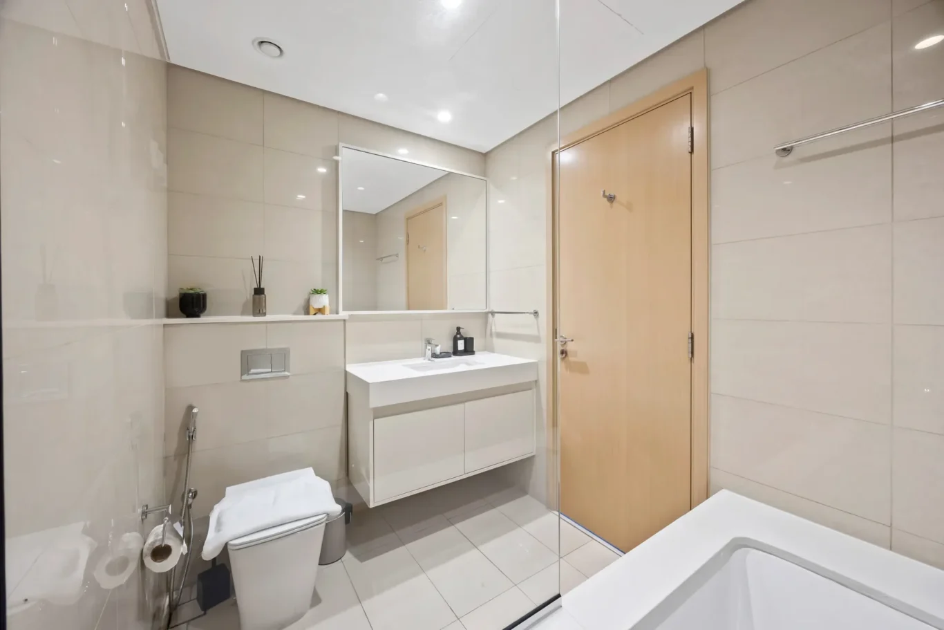 Modern beige-tiled bathroom with a white wall-mounted vanity, large mirror, and glass shower enclosure beside a wooden door.