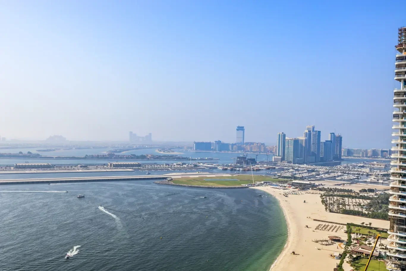 Aerial view of a coastal city: a curved sandy beach beside a calm bay, with a marina and cluster of modern high-rise buildings in the distance.
