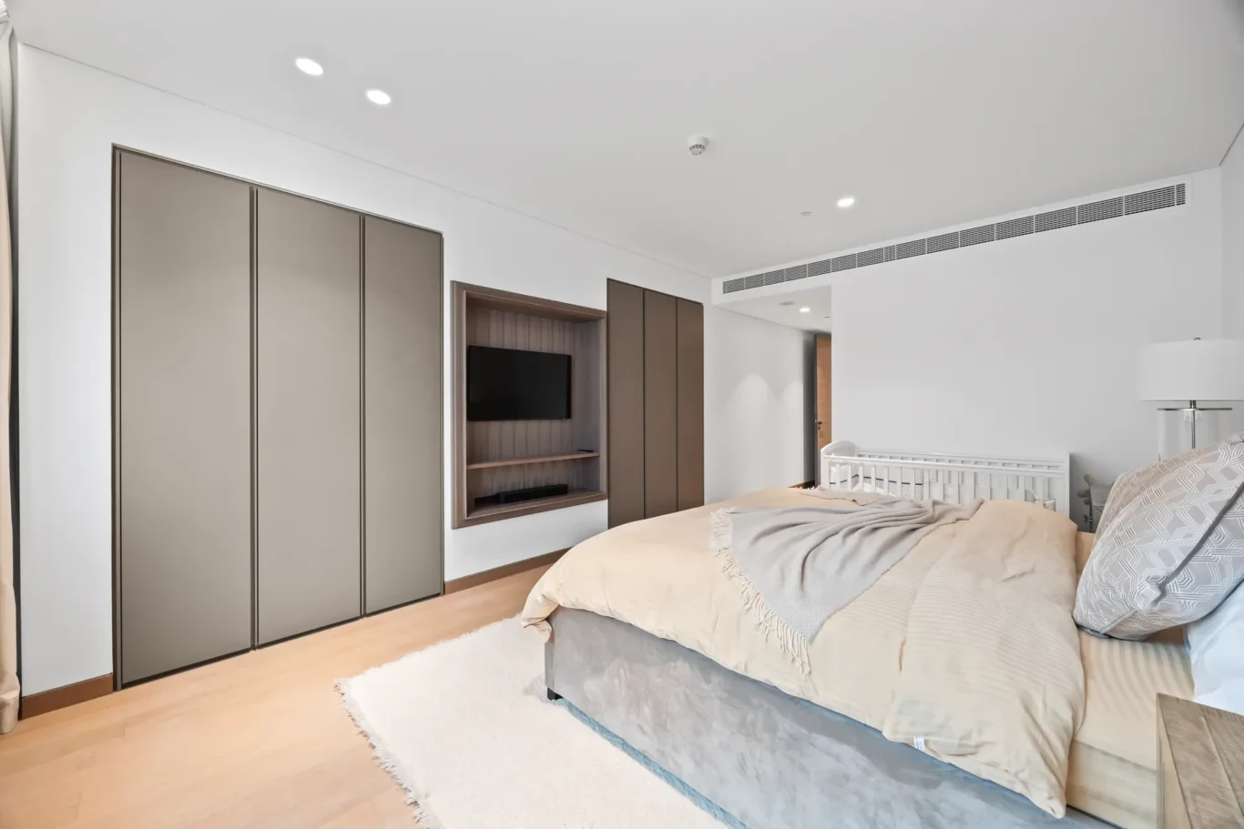 Modern bedroom with large beige built-in wardrobes, a wood alcove housing a wall-mounted TV, and a bed with beige bedding on a white rug. A white crib sits beside the bed.