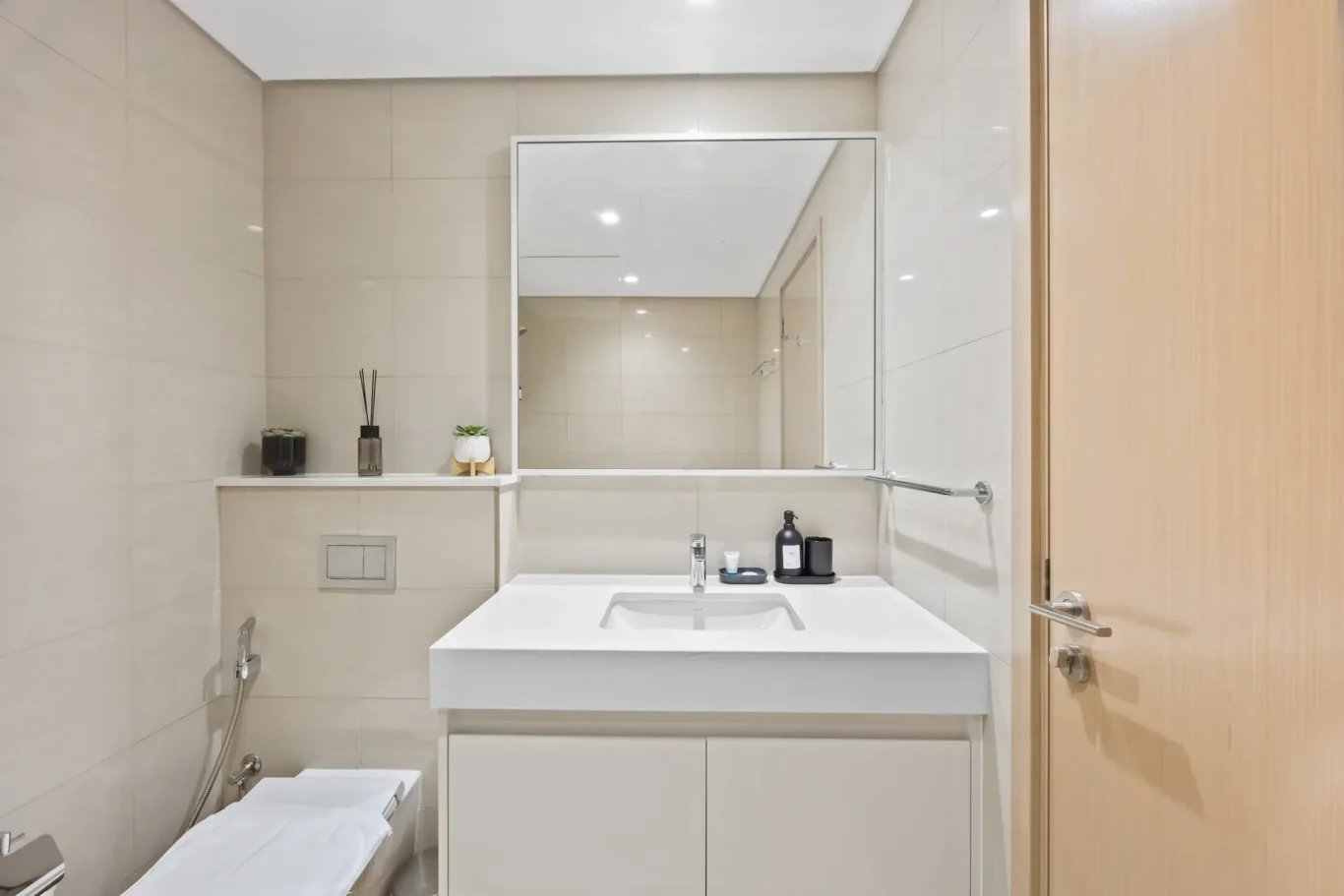 Modern beige-tiled bathroom with a white vanity, large mirror, and wooden door on the right.