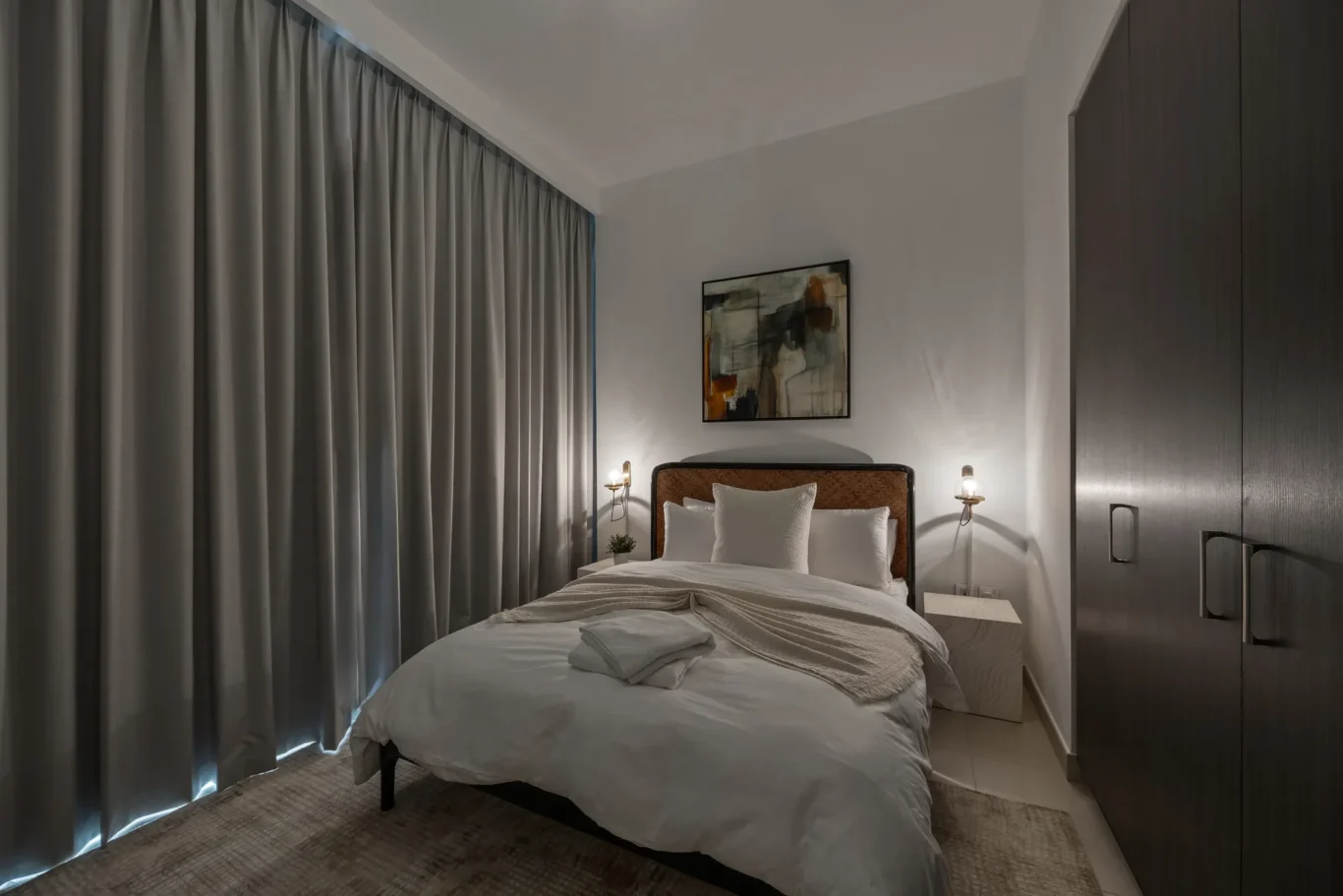 Hotel bedroom with double bed, gray curtains, abstract painting, and warm wall lamps above each side of the bed.