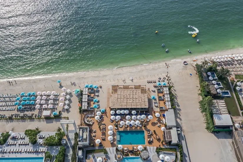 Aerial view of a beach resort: turquoise water, sandy shore, and a large pool area with blue umbrellas and loungers surrounding buildings along the coast.
