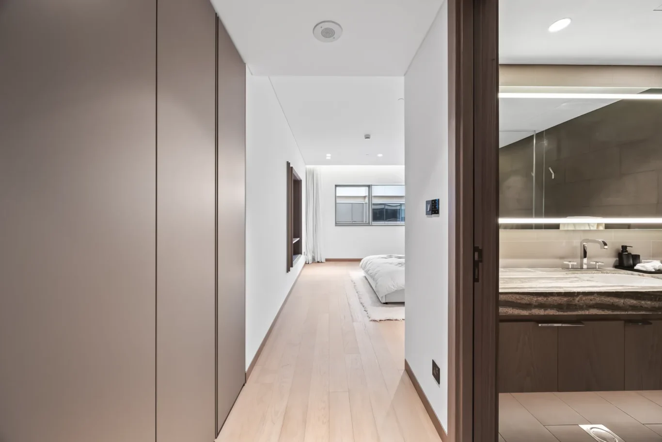 Minimalist hallway opening onto a bright bedroom with a white bed, light flooring, and tall brown wardrobes on the left; bathroom area visible to the right.