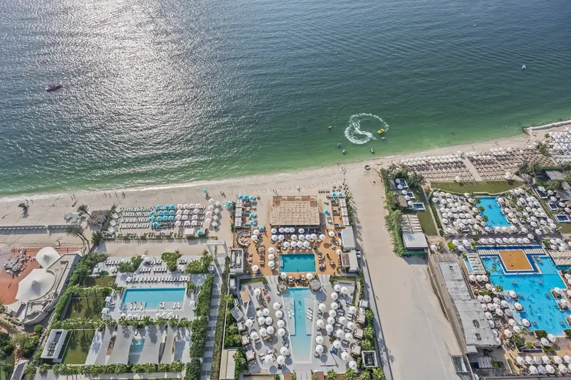 Aerial view of a seaside resort: sandy beach, turquoise water, and numerous pool areas with sun loungers and cabanas surrounded by buildings.